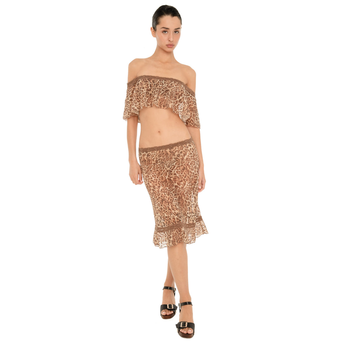 Full body front view of a model wearing the Liulu Leopard Top, an off shoulder cropped top with a brown leopard print, lace trimmed neckline and flowing ruffle overlay, styled with a matching leopard print midi skirt and black heels