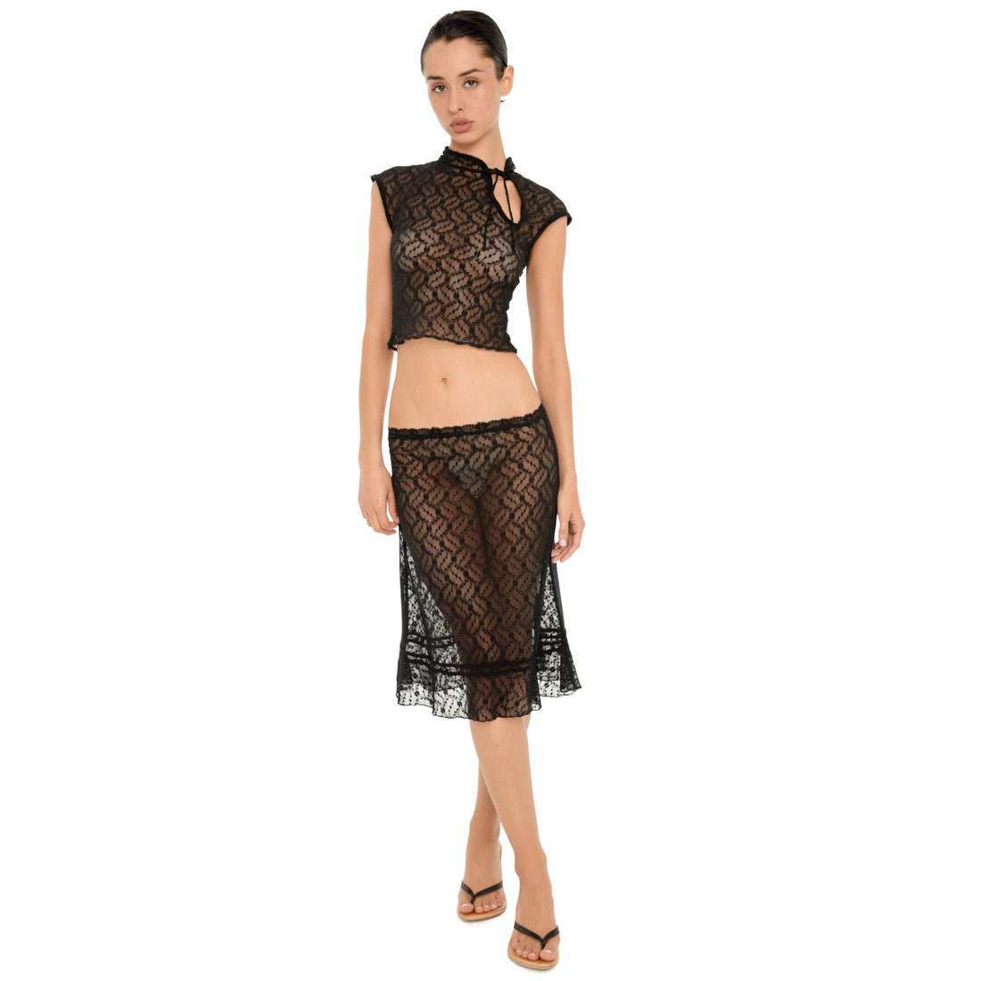 Full-length front view of a model wearing a sheer black floral lace midi skirt with a low-rise waist and ruffled hem, styled with a matching black lace top and black heeled sandals. **black