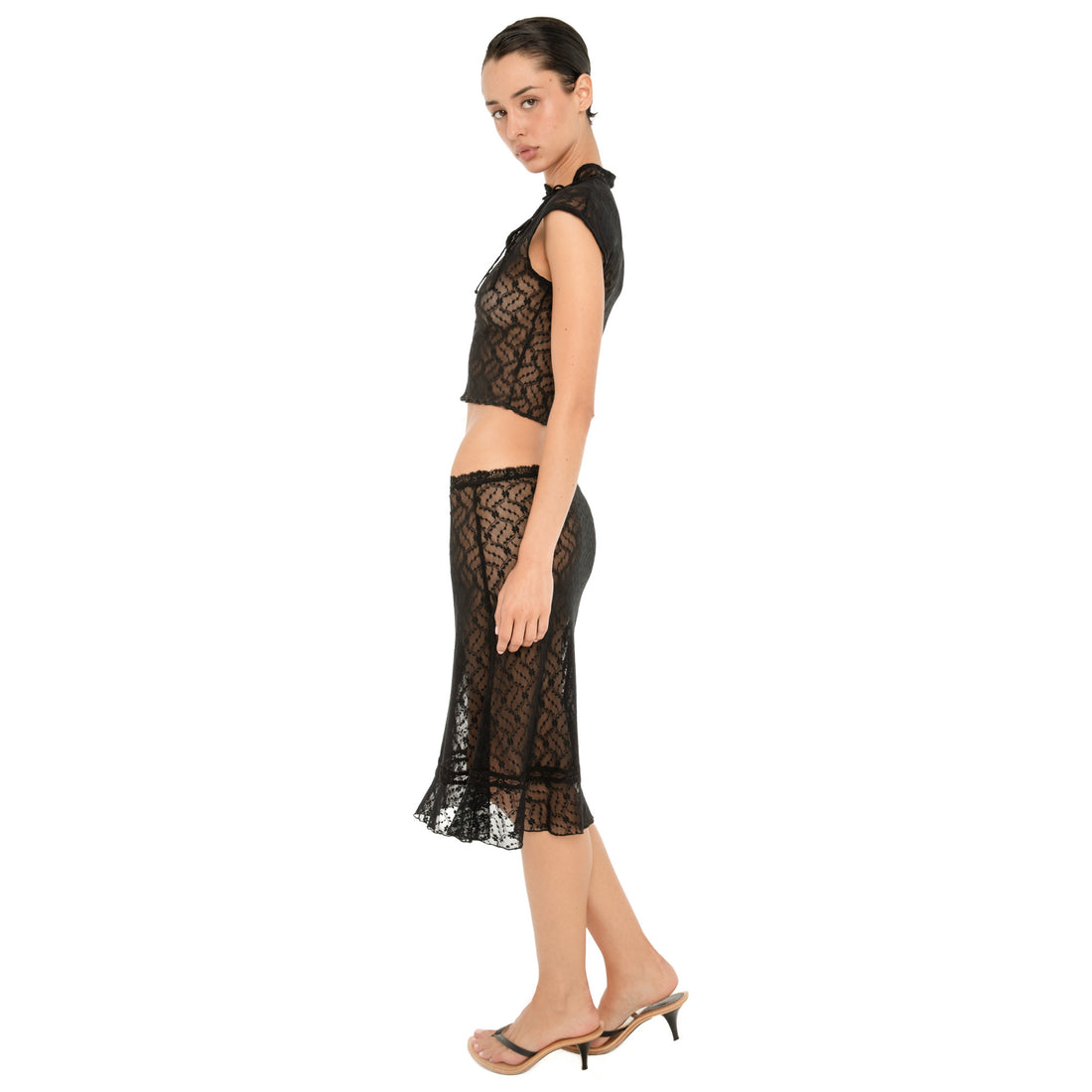 Full-length side view of a model wearing the sheer black floral lace Liulu Skirt, showing its low-rise waist, sheer lace construction, fitted hips, and flared ruffled hem. **black