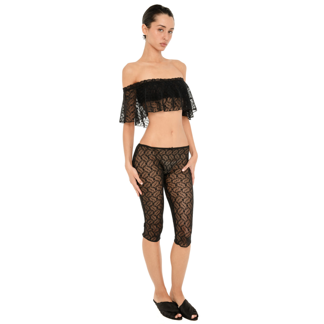 Front view of a model wearing a sheer black floral lace off-the-shoulder cropped top with a ruffled overlay and matching low-rise fitted lace capri pants against a white background. **black