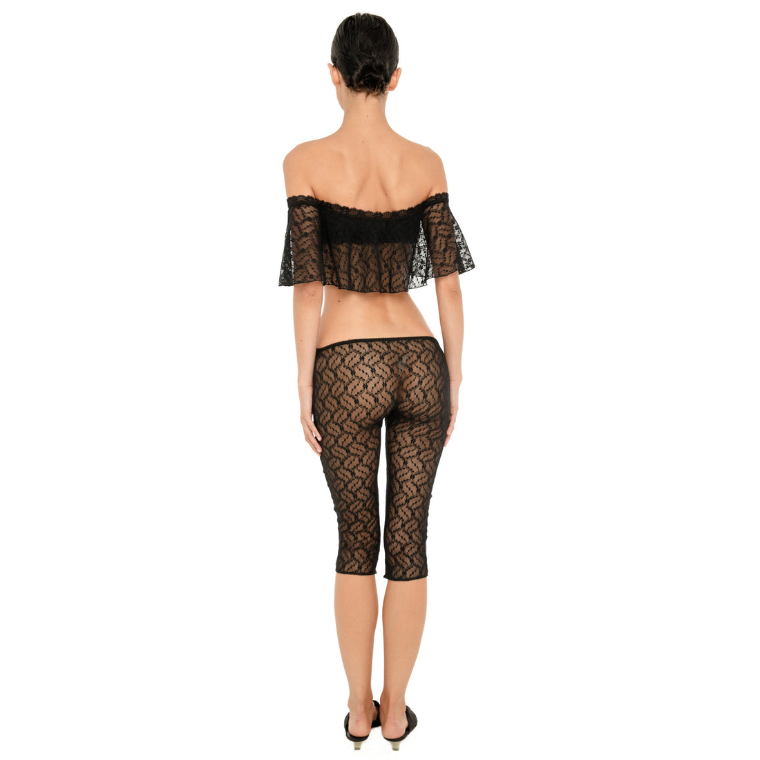 Back view of a model wearing the sheer black floral lace off-the-shoulder cropped top and matching low-rise fitted capri pants against a white background. **black
