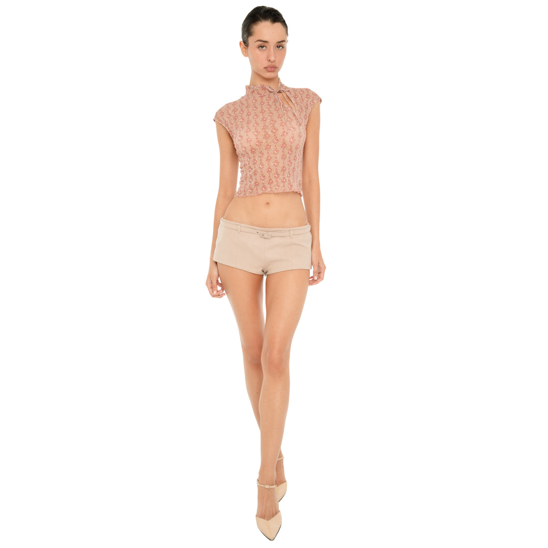 Full-length front view of model wearing the ARE YOU AM I Pika Short in Almond, a low-rise micro short with a slim matching belt, styled with a floral top and pointed heels. **almond