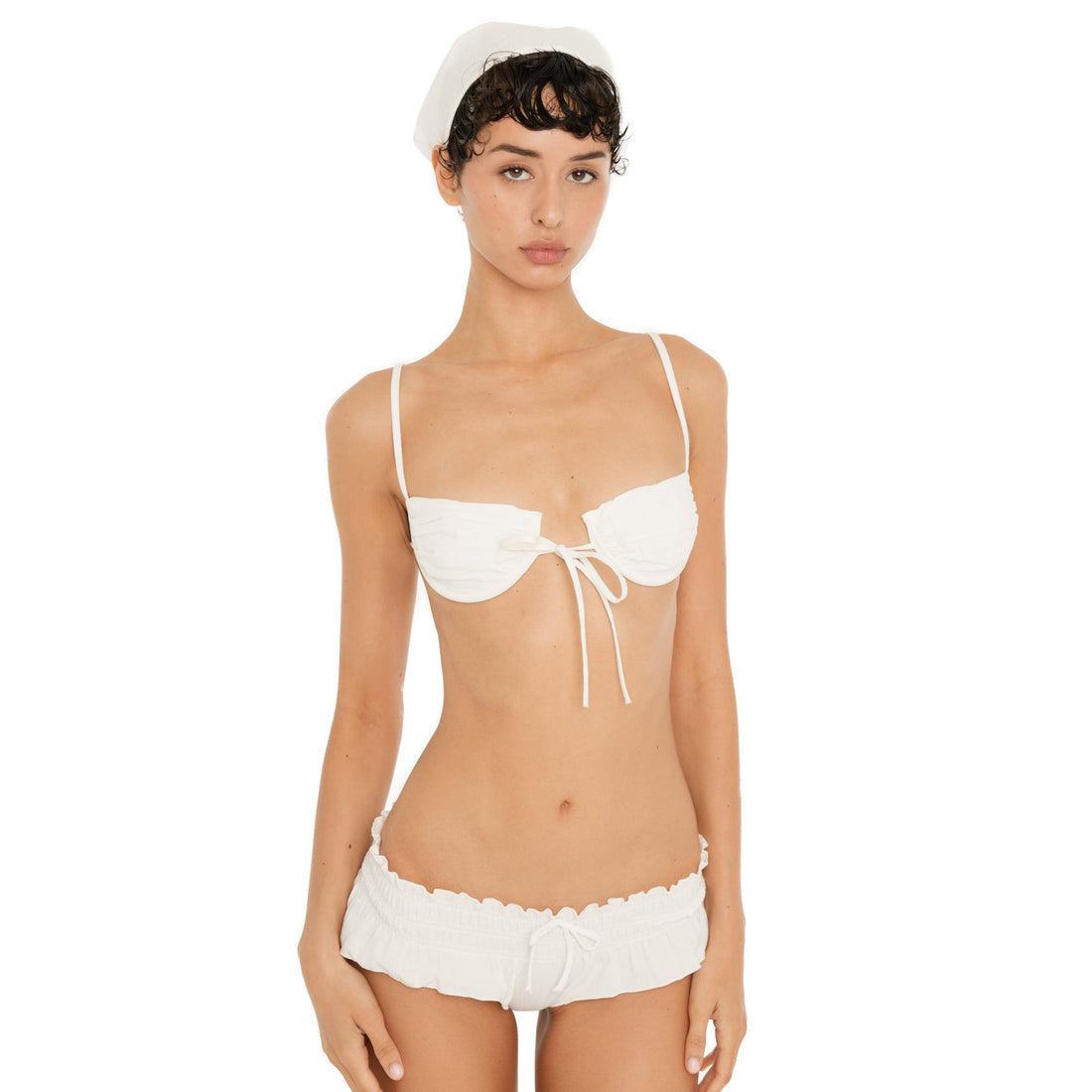 Model wearing the natural cotton Liette Headkerchief tied over her dark hair, styled with a white ruched bra top and low-rise shorts.