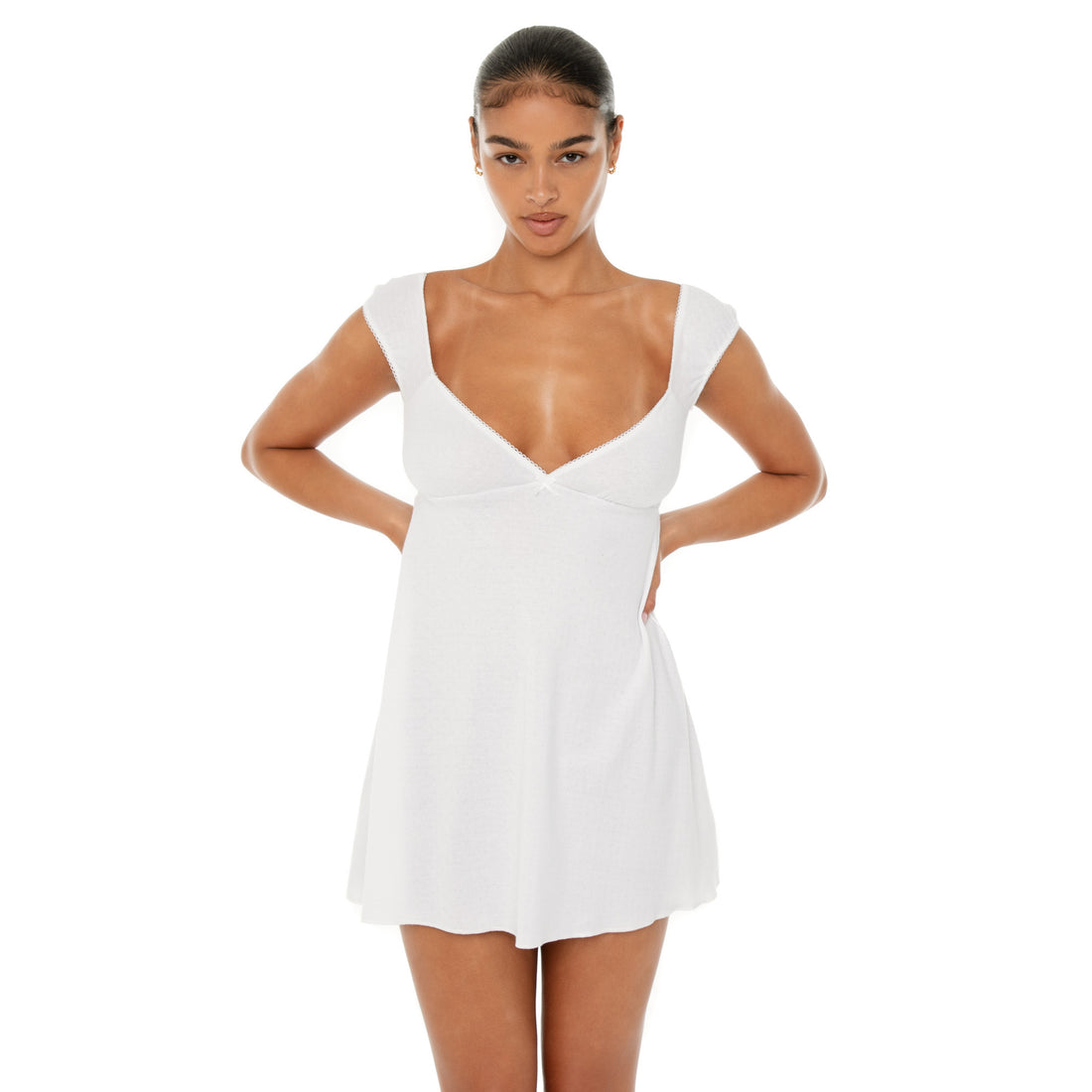 Model wearing the Eveli Dress — white cotton mini with cap sleeves and deep scoop neckline, finished with delicate scalloped trim and subtle sheer texture.