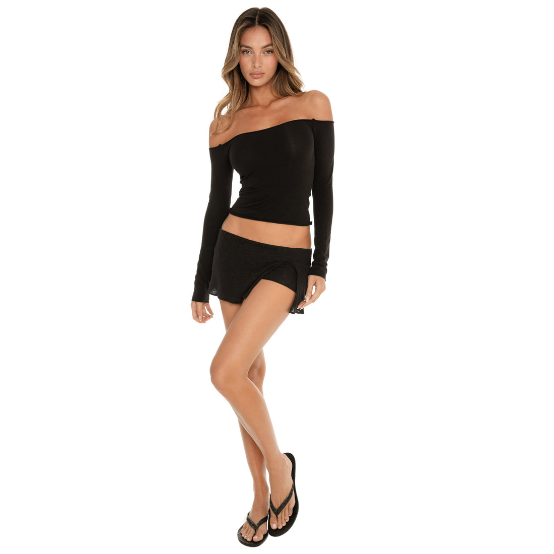Model standing in the Anouck Top in black, off shoulder long sleeve silhouette paired with a black low rise skort, full body front pose, wearing black flip flops **black