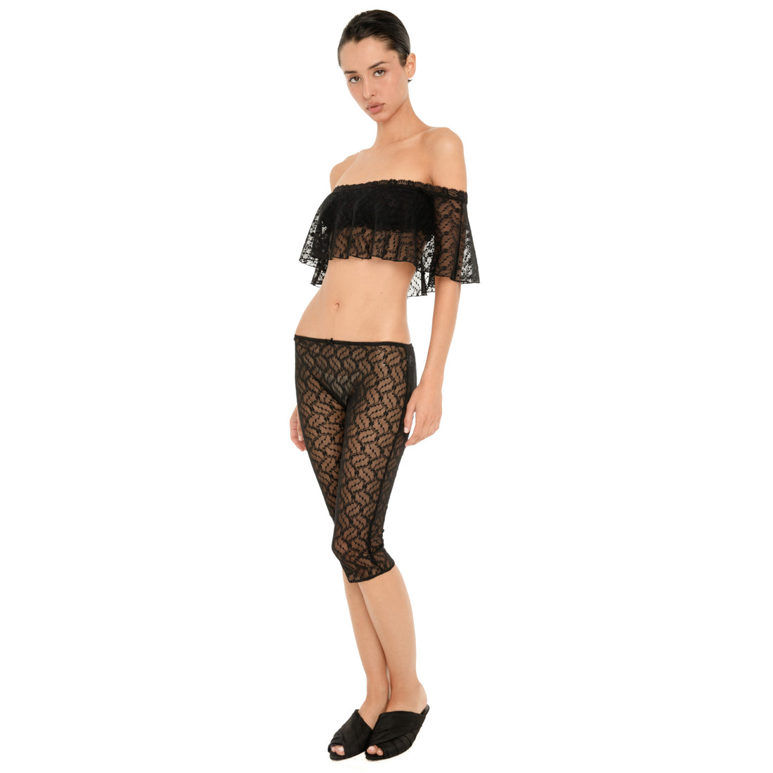 Model wearing a sheer black floral lace off-the-shoulder cropped top with a ruffled overlay and matching fitted lace capri pants, shown in a three-quarter front view against a white background. **black