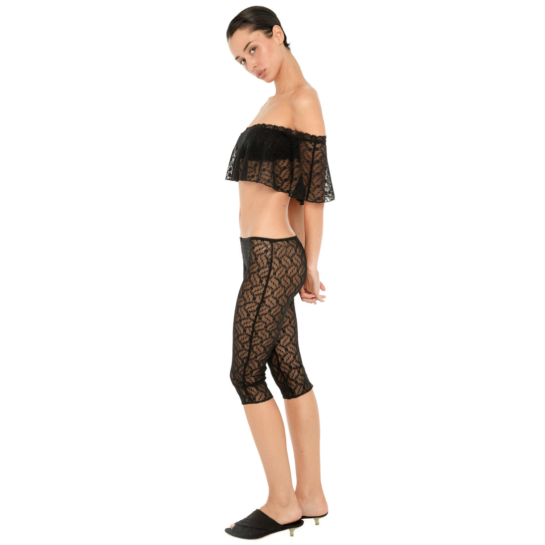 Side view of a model wearing the sheer black floral lace cropped top and matching fitted capri pants, showing the lace pattern and side silhouette against a white background. **black