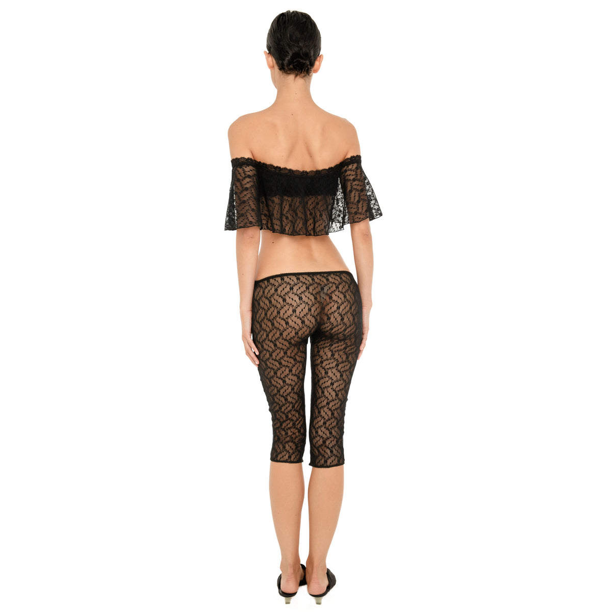 Back view of a model wearing the sheer black floral lace off-the-shoulder cropped top and matching low-rise fitted capri pants against a white background. **black