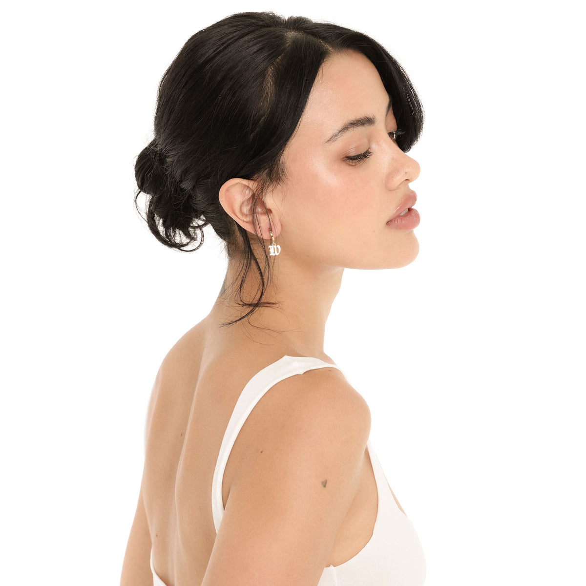 Side profile of a model wearing the Nimi Earring in W, a small gold huggie hoop with a dangling gothic-script letter W charm, styled with her hair in a low bun