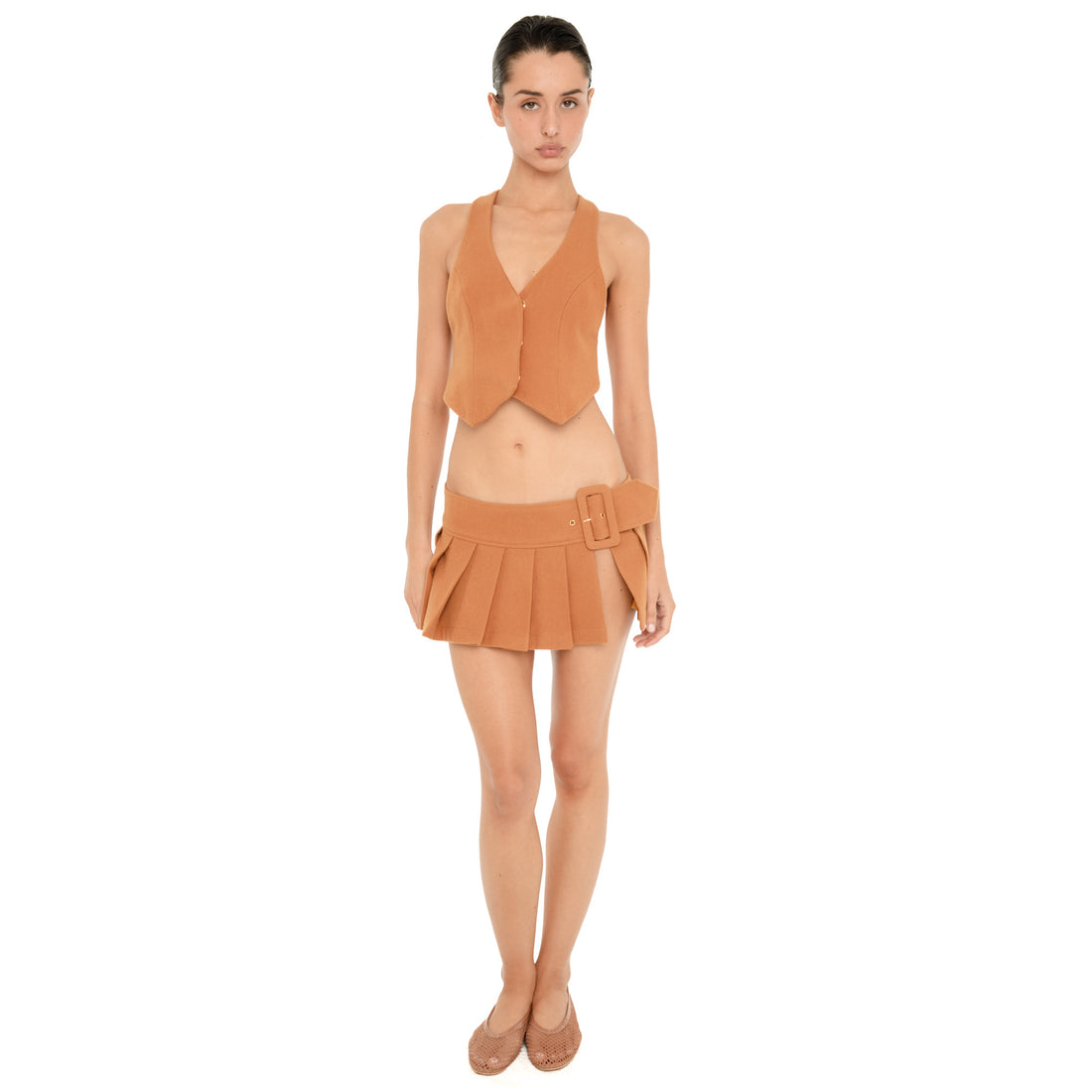 Full-length front view of model wearing the ARE YOU AM I Pau Vest in Saffron, a fitted cropped vest with a deep V neckline and pointed hem, styled with a matching pleated mini skirt. **saffron