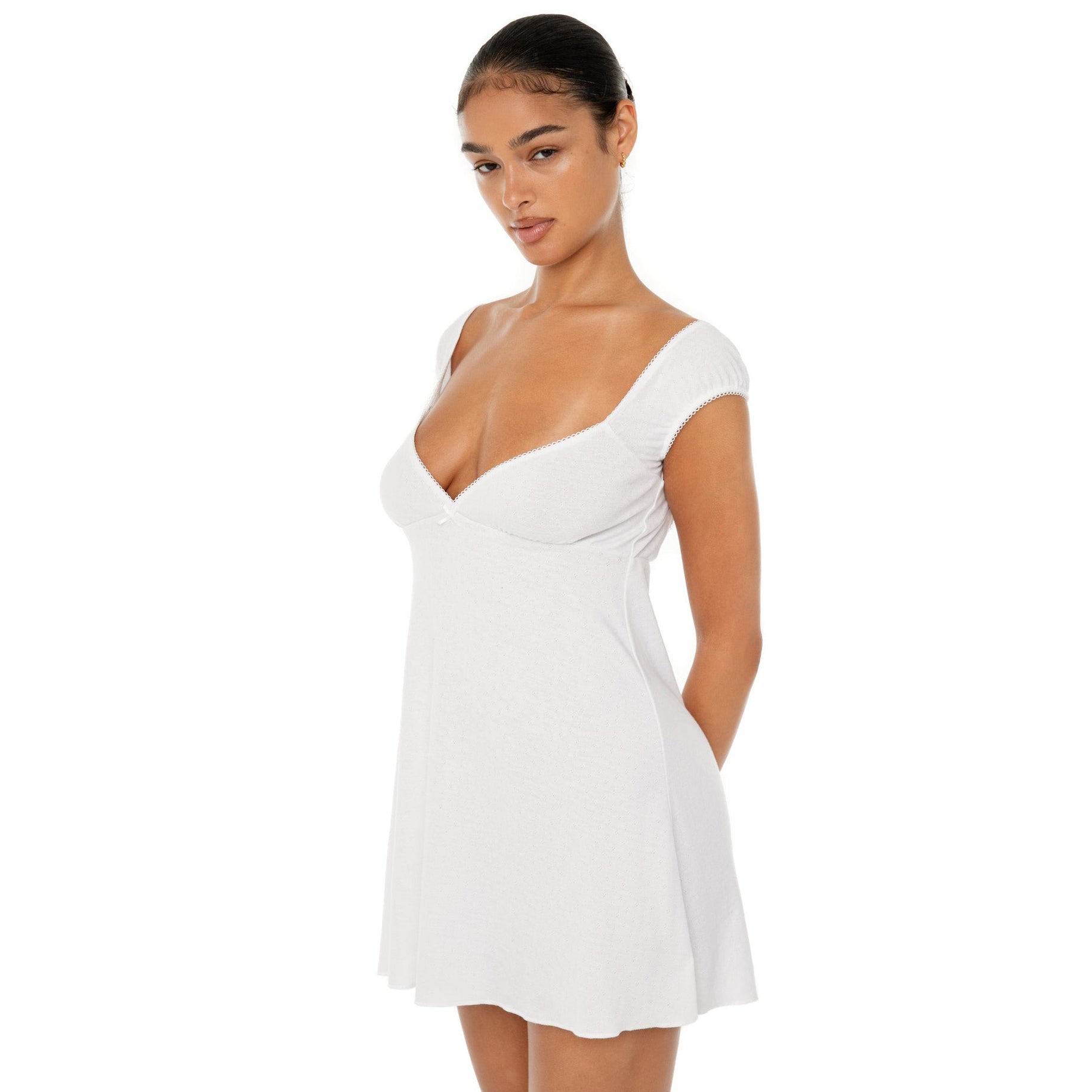Model wearing the Eveli Dress — white cotton mini with cap sleeves and deep scoop neckline, finished with delicate scalloped trim and subtle sheer texture.