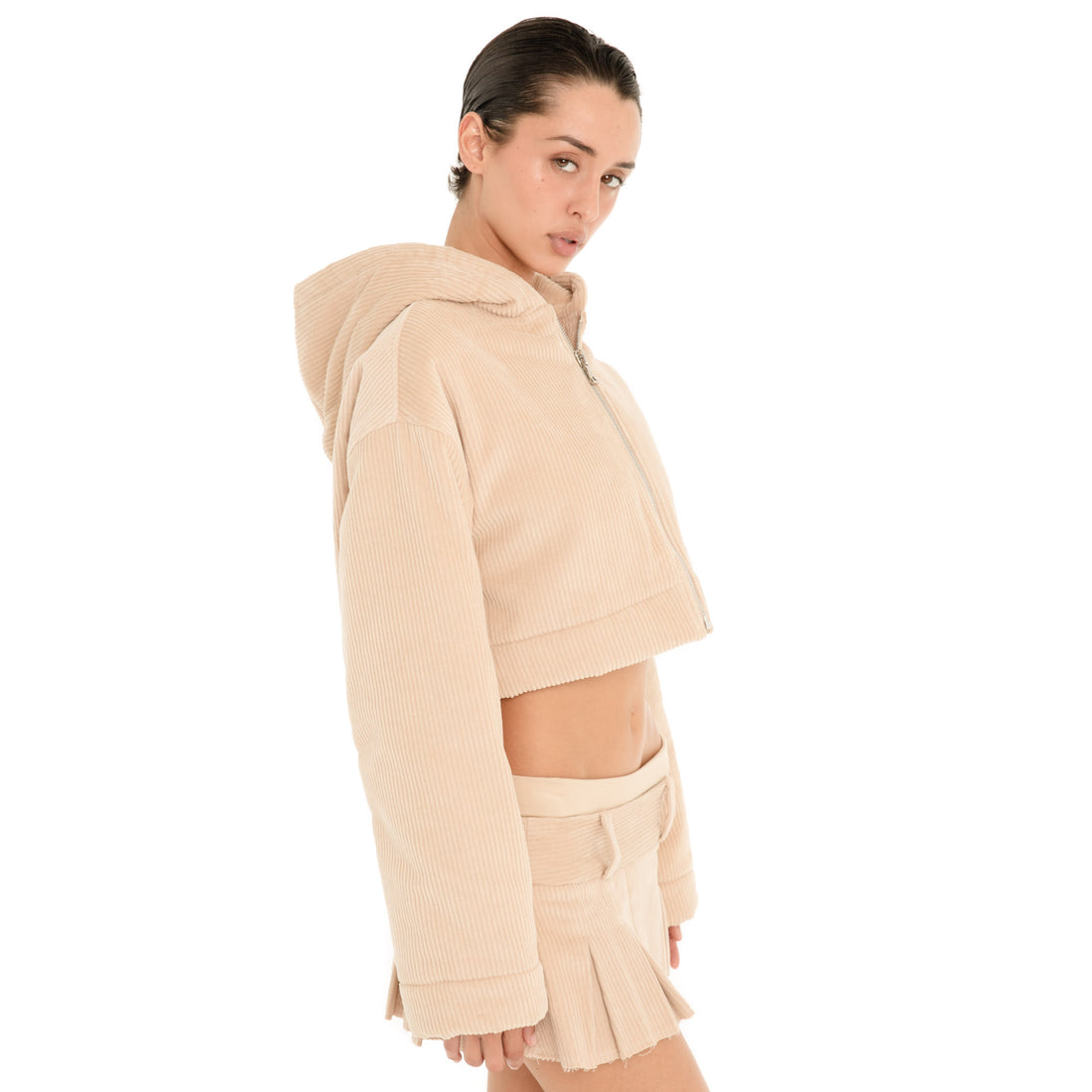 Three-quarter view of model wearing the Sfera Jacket in Sand, a cropped ribbed hooded jacket with a front zipper and long relaxed sleeves, styled with a pleated mini skirt. **sand