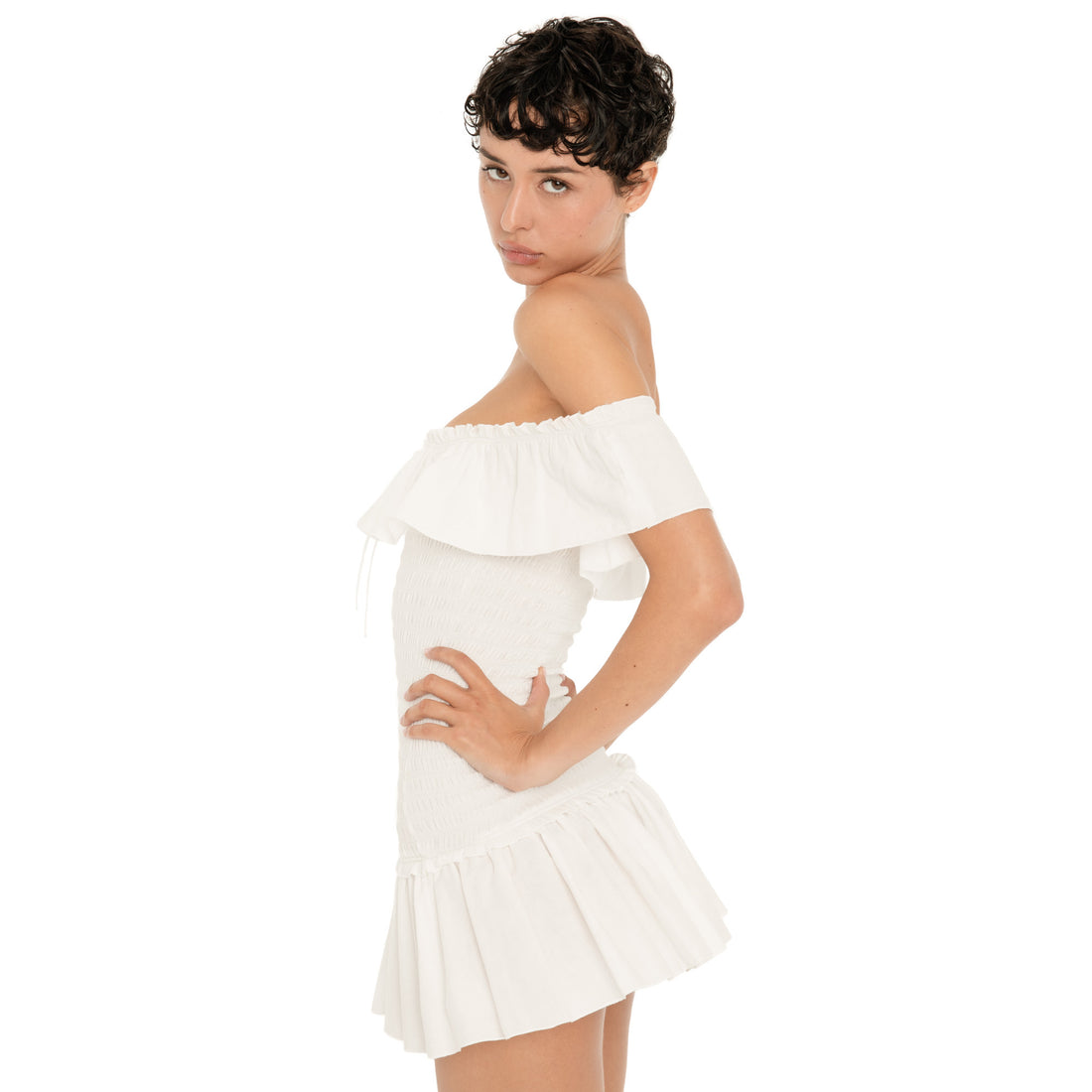 Three quarter side view of the white Zouette mini dress, showing the off shoulder ruffle, fitted smocked bodice, and flared pleated mini skirt