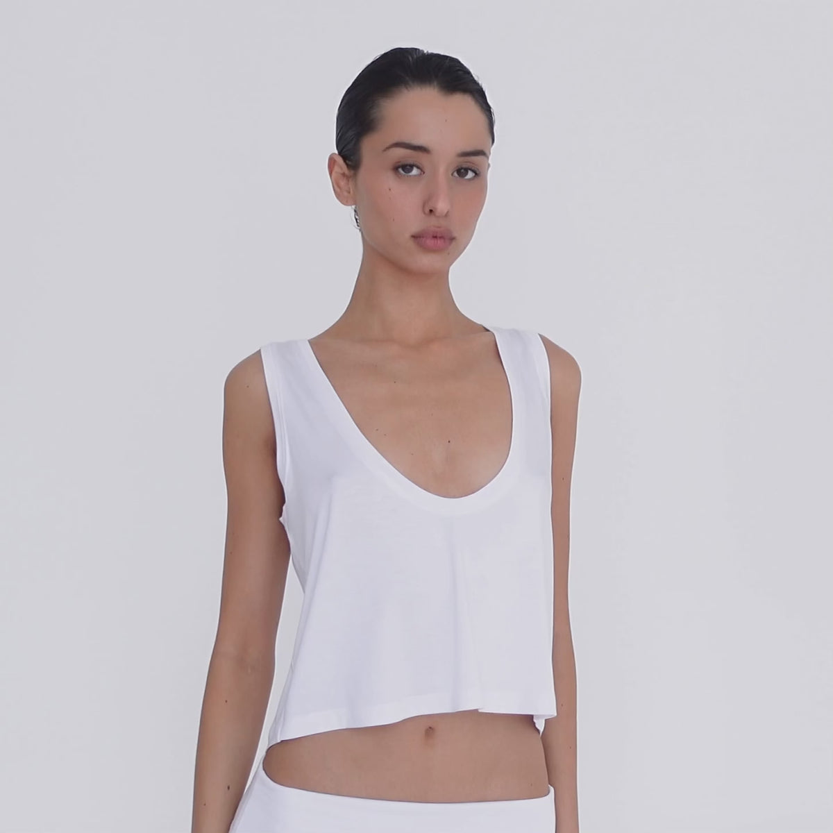 Video: Model moves to show the white Kasia Top from multiple angles, highlighting its deep scoop neckline, wide straps, cropped length and fluid drape. **white