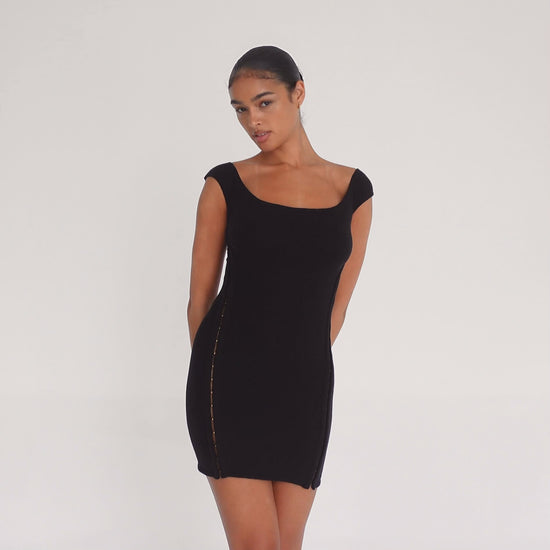 Model wearing the black Nikisa Dress — off-shoulder ribbed mini with gold hook-and-eye closures running down each side, hugging the body in a sculpted silhouette.**black
