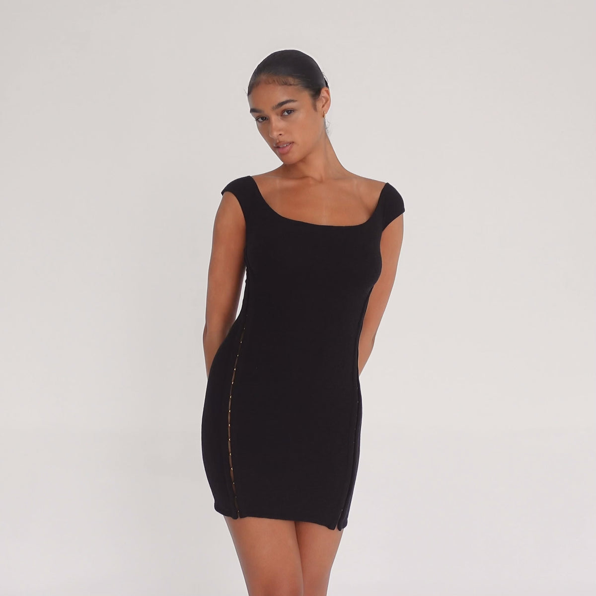 Model wearing the black Nikisa Dress — off-shoulder ribbed mini with gold hook-and-eye closures running down each side, hugging the body in a sculpted silhouette.**black