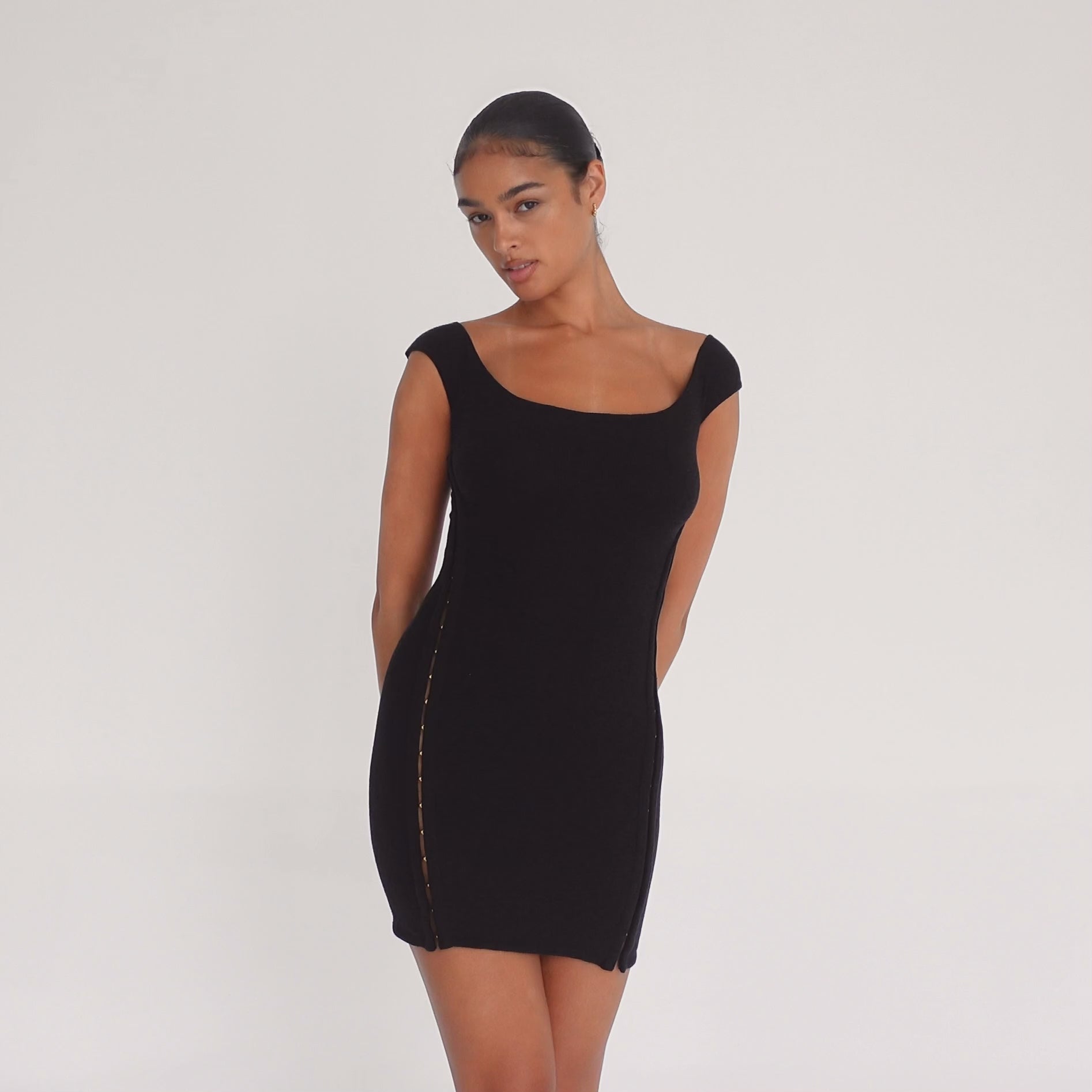 Model wearing the black Nikisa Dress — off-shoulder ribbed mini with gold hook-and-eye closures running down each side, hugging the body in a sculpted silhouette.**black
