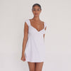 Model wearing the Eveli Dress — white cotton mini with cap sleeves and deep scoop neckline, finished with delicate scalloped trim and subtle sheer texture.