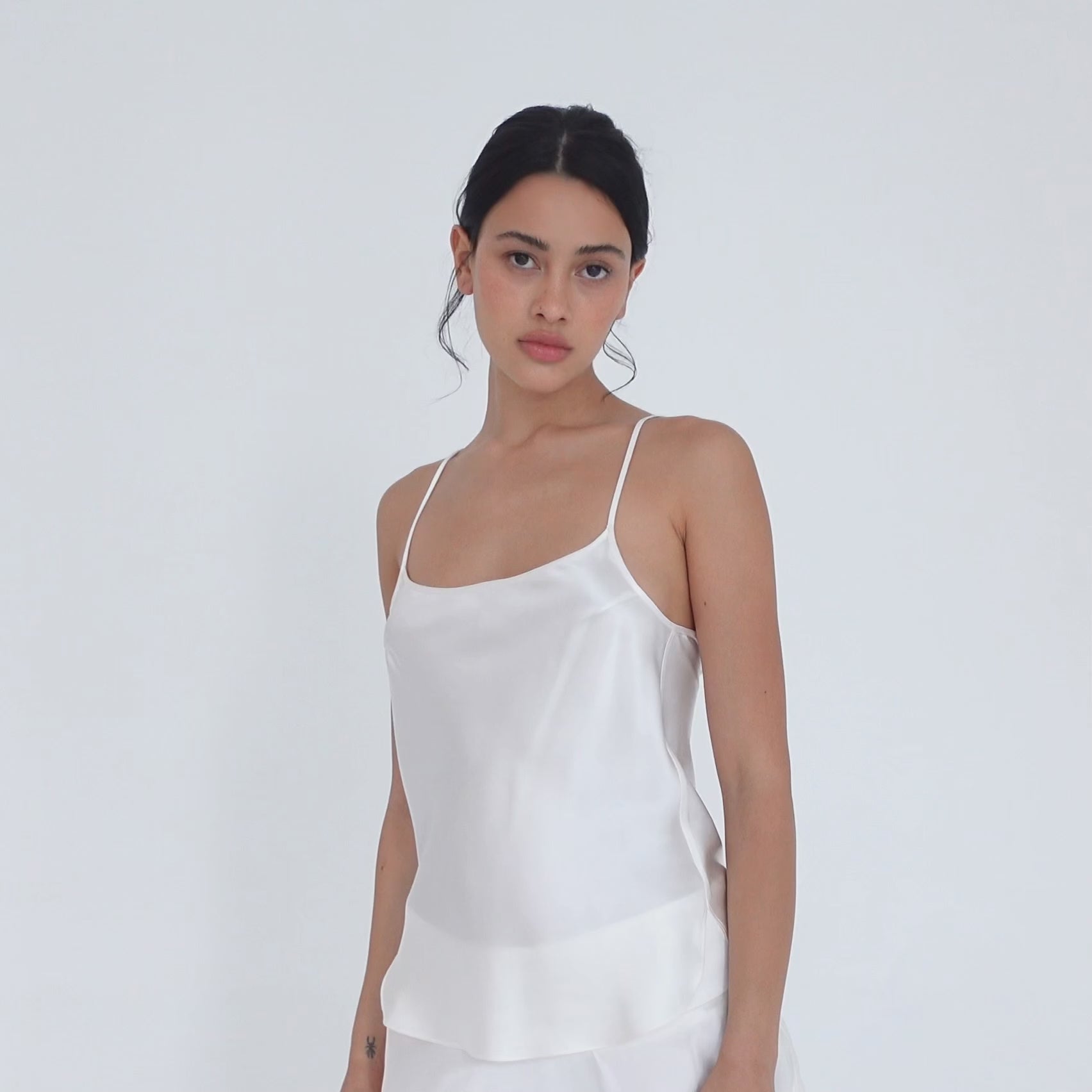 Model turning slowly in the white silk Syra Camisole, showing the scoop neckline from the front, the curved side hem and the low scoop back. **white