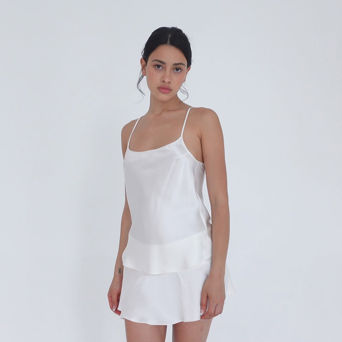 Model turning slowly in the white silk Syra Skirt and Syra Camisole, showing the low-rise waistband, the flared mini hem from the front and side, and the back. **white