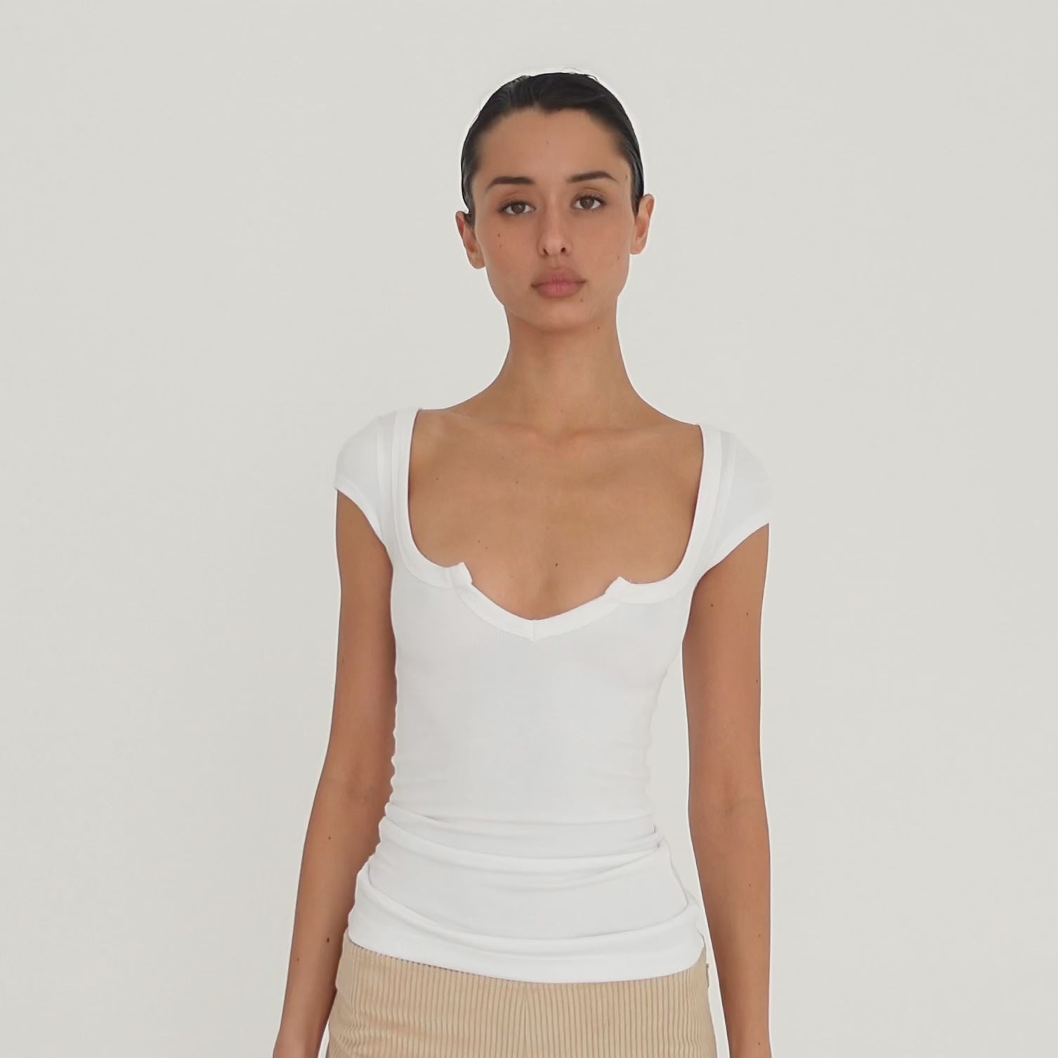Video: Model moves to show the white Impy Tee from multiple angles, highlighting its fitted ribbed silhouette, cap sleeves and sculpted notched neckline. **white